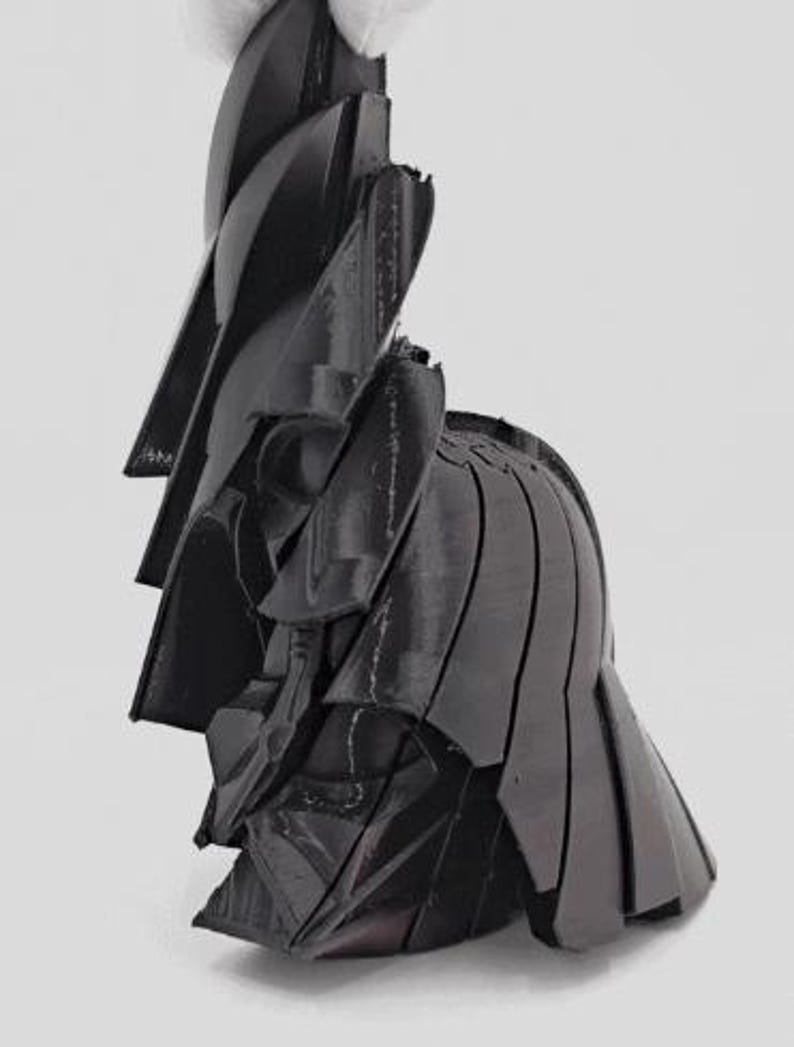 Pode incluir: Uma escultura abstrata preta com um design em camadas. A escultura parece ser feita de v&aacute;rias pe&ccedil;as sobrepostas, criando uma forma texturizada e din&acirc;mica. O objeto &eacute; segurado por uma m&atilde;o enluvada contra um fundo branco.