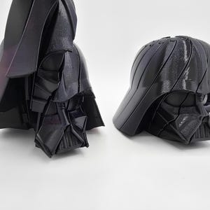 Pode incluir: Dois modelos de capacete Darth Vader pretos impressos em 3D. Um modelo &eacute; um capacete completo com capa, e o outro &eacute; um capacete estilo bon&eacute; de beisebol. Os capacetes s&atilde;o brilhantes e t&ecirc;m um design angular detalhado.