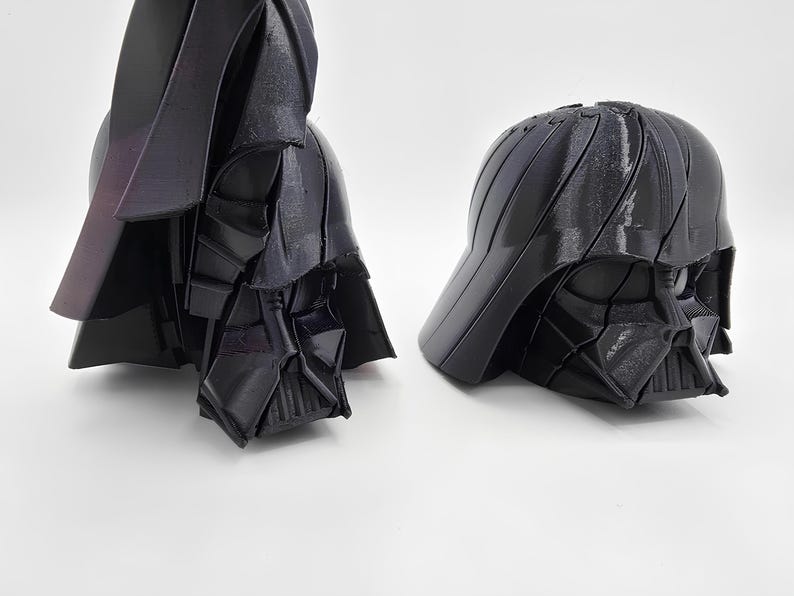 Pode incluir: Dois modelos de capacete Darth Vader pretos impressos em 3D. Um modelo &eacute; um capacete completo com capa, e o outro &eacute; um capacete estilo bon&eacute; de beisebol. Os capacetes s&atilde;o brilhantes e t&ecirc;m um design angular detalhado.