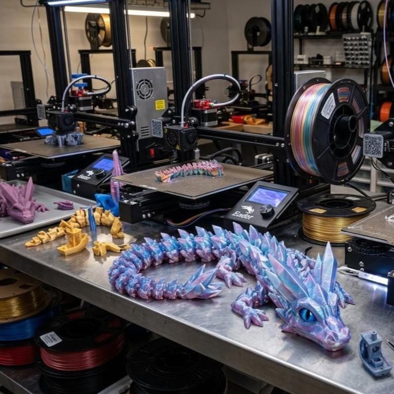 May include: A 3D printed articulated dragon with iridescent blue and purple scales and large blue eyes, resting on a metal surface. Several 3D printers are visible in the background with spools of multi-colored filament and other printed parts.