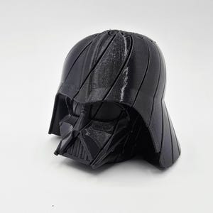Pode incluir: Um capacete preto de Darth Vader, um personagem reconhec&iacute;vel da franquia Star Wars. O capacete &eacute; feito de um material escuro com uma superf&iacute;cie texturizada e est&aacute; sobre um fundo branco. O design do capacete &eacute; detalhado e ic&ocirc;nico.