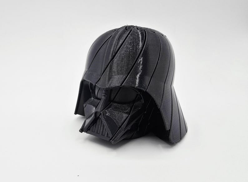 Pode incluir: Um capacete preto de Darth Vader, um personagem reconhec&iacute;vel da franquia Star Wars. O capacete &eacute; feito de um material escuro com uma superf&iacute;cie texturizada e est&aacute; sobre um fundo branco. O design do capacete &eacute; detalhado e ic&ocirc;nico.