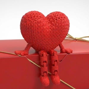 Pode incluir: Uma figura de coração vermelho tricotado com pernas e braços articulados senta-se em cima de uma caixa de presente vermelha amarrada com uma fita dourada. A textura do coração é visível, e o design é caprichoso e romântico.