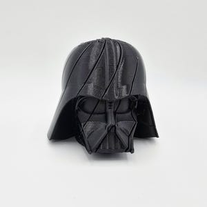 Pode incluir: Um capacete preto de Darth Vader impresso em 3D, um personagem da franquia Star Wars. O capacete apresenta detalhes faciais distintos e uma superf&iacute;cie texturizada. A imagem &eacute; contra um fundo branco liso.