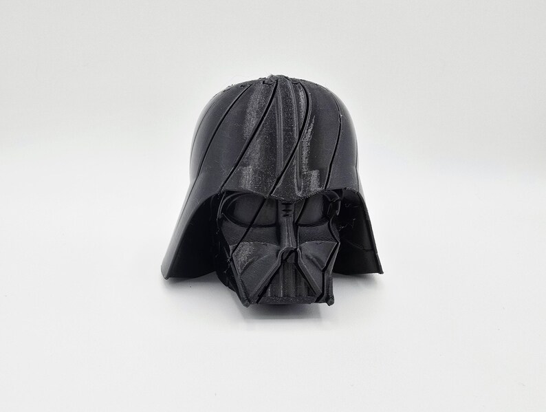Pode incluir: Um capacete preto de Darth Vader impresso em 3D, um personagem da franquia Star Wars. O capacete apresenta detalhes faciais distintos e uma superf&iacute;cie texturizada. A imagem &eacute; contra um fundo branco liso.