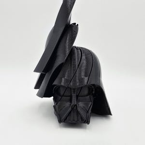 Pode incluir: Um capacete preto de Darth Vader impresso em 3D, com o ic&oacute;nico design de m&aacute;scara e capacete. O capacete est&aacute; angulado, mostrando os detalhes da m&aacute;scara facial e a parte superior do capacete. A imagem est&aacute; sobre um fundo branco.