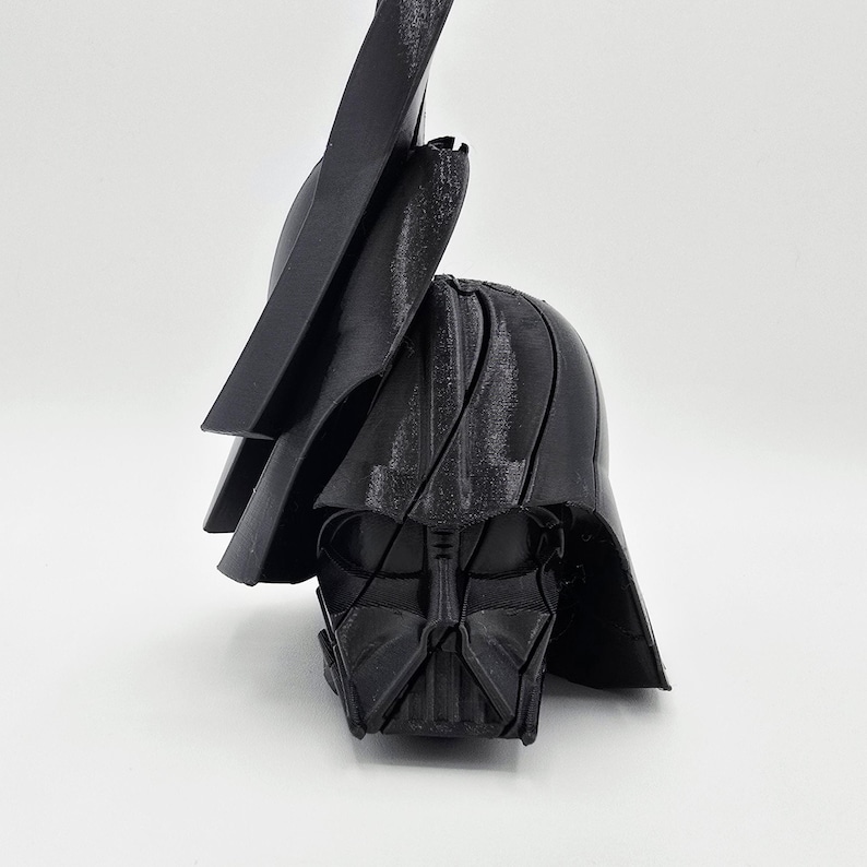 Pode incluir: Um capacete preto de Darth Vader impresso em 3D, com o ic&oacute;nico design de m&aacute;scara e capacete. O capacete est&aacute; angulado, mostrando os detalhes da m&aacute;scara facial e a parte superior do capacete. A imagem est&aacute; sobre um fundo branco.