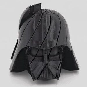 Pode incluir: Um capacete preto de Darth Vader impresso em 3D, apresentando o design ic&ocirc;nico da m&aacute;scara e do capacete. O capacete &eacute; dividido em v&aacute;rias se&ccedil;&otilde;es, com linhas vis&iacute;veis. A imagem &eacute; tirada contra um fundo liso e claro.