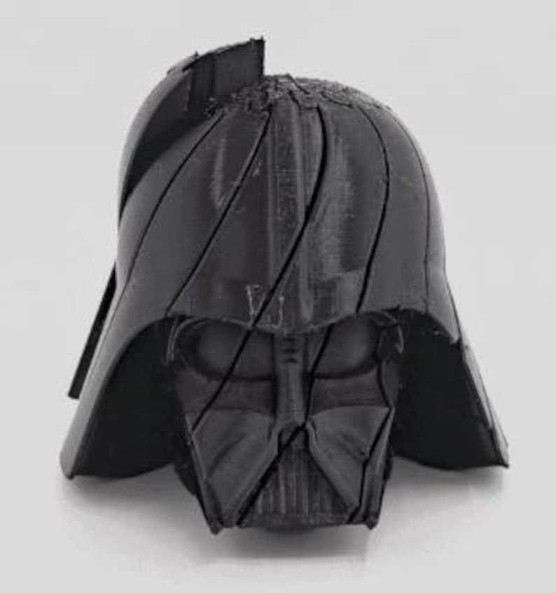 Pode incluir: Um capacete preto de Darth Vader impresso em 3D, apresentando o design ic&ocirc;nico da m&aacute;scara e do capacete. O capacete &eacute; dividido em v&aacute;rias se&ccedil;&otilde;es, com linhas vis&iacute;veis. A imagem &eacute; tirada contra um fundo liso e claro.