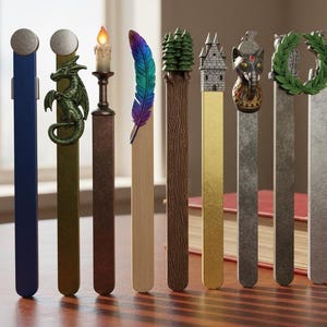 Op de afbeelding: Een verzameling decoratieve bladwijzers met verschillende ontwerpen. De bladwijzers hebben een reeks versieringen, waaronder een draak, een kaars, een veer, een boom, een kasteel, een olifant, een krans en een vos. Elke bladwijzer heeft een andere kleur en stijl.