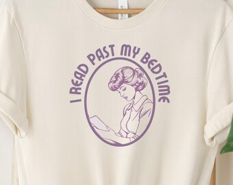 Könnte beinhalten: Ein cremefarbenes T-Shirt mit einer lilafarbenen Grafik einer Frau, die ein Buch liest. Die Grafik befindet sich in einem Kreis mit dem Text "I read past my bedtime".