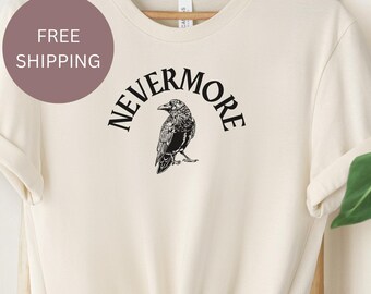 May include: A cream-colored short-sleeve t-shirt featuring a black graphic of a raven centered on the chest. The word NEVERMORE is printed in a black arched font above the bird illustration. The shirt has a crew neckline and rolled sleeves.