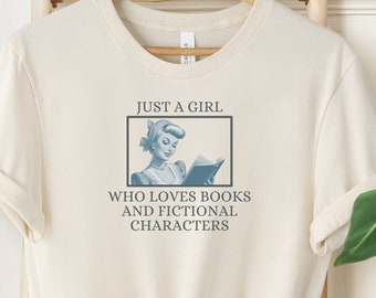 Op de afbeelding: Een crèmekleurig T-shirt met een afbeelding van een vrouw die een boek leest. De tekst op het shirt luidt "Just a girl who loves books and fictional characters."