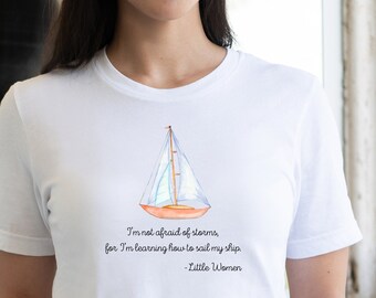 Può includere: Maglietta bianca con un'illustrazione ad acquerello di una barca a vela e una citazione di Piccole donne: "Non ho paura delle tempeste, perché sto imparando a navigare la mia nave."