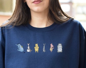 Op de afbeelding: Een donkerblauwe trui met een afbeelding van Winnie de Poeh en zijn vrienden. De personages staan in een rij en omvatten Biggetje, Eeyore, Teigetje, Uil en Konijn.