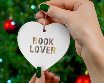 Puede incluir: Adorno blanco en forma de corazón con el texto "BOOK LOVER" en letras de colores. El adorno está siendo sostenido por una mano con esmalte de uñas verde sobre un fondo de árbol de Navidad verde.