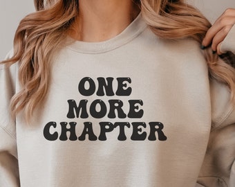 May include: A light beige sweatshirt with the text "ONE MORE CHAPTER" printed in black in a retro font.