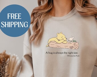 Op de afbeelding: Een beige sweatshirt met een illustratie van Winnie de Poeh en Biggetje, met de quote "Een knuffel is altijd de juiste maat. -Winnie de Poeh".