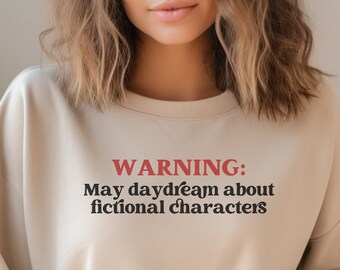 Könnte beinhalten: Ein beiger Pullover mit dem Text "WARNING: May daydream about fictional characters" in Schwarz und Rot.