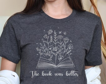 Puede incluir: Una camiseta gris oscuro con un contorno blanco de un libro abierto con flores que salen de él. El texto "The book was better" está impreso debajo del libro.