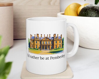 May include: White ceramic mug featuring a detailed illustration of a stately yellow building with a blue roof. The mug has the text "I'd rather be at Pemberley" printed in black.