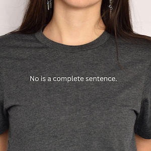 May include: Dark gray crew neck t-shirt with the text "No is a complete sentence" in white. The shirt is made of a soft, heathered fabric. The image shows the upper torso of a person wearing the shirt.
