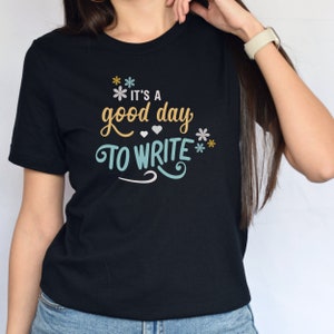 May include: Black t-shirt with a white, blue, and yellow graphic that says "It's a good day to write" with flowers and hearts.
