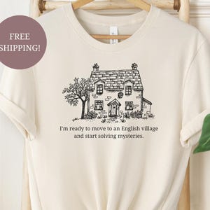 May include: Cream-colored t-shirt featuring a black and white illustration of a quaint English cottage with a tree. The text below the image reads, "I'm ready to move to an English village and start solving mysteries."