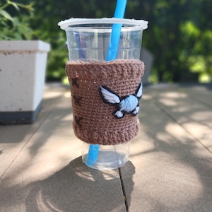 May include: A clear plastic cup with a blue straw, encased in a brown crocheted cozy. The cozy features a white and blue winged design. The cup sits on a wooden surface, with a blurred green background.