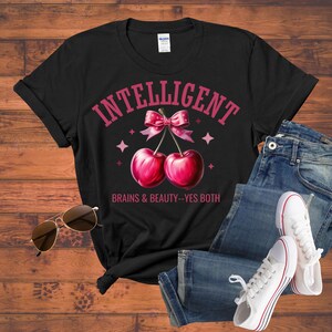 May include: Black t-shirt with a pink graphic of two cherries tied with a bow, the word "INTELLIGENT" above, and the text "BRAINS & BEAUTY--YES BOTH" below. Sunglasses, jeans, and white sneakers are also in the image.