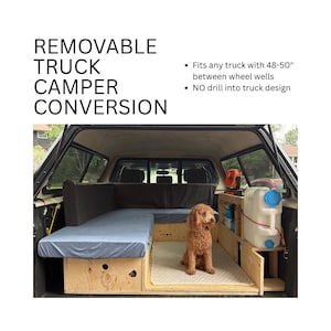 May include: A truck camper conversion with a wooden frame, a blue mattress, and storage compartments. The text reads "REMOVABLE TRUCK CAMPER CONVERSION." It fits trucks with 122-127 cm between wheel wells. A dog sits inside the camper.