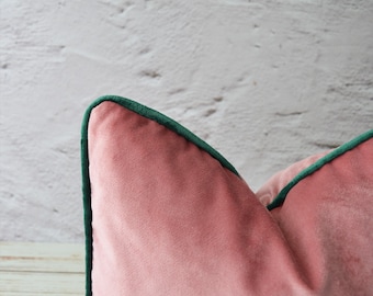 Puede incluir: Un cojín decorativo de terciopelo rosa empolvado suave con un borde ribeteado en color verde bosque oscuro. El cojín tiene una textura afelpada y lisa, con una costura en la esquina que resalta el ribete verde vibrante sobre la tela rosa.