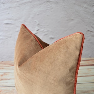 May include: A square throw pillow made of tan velvet fabric. The pillow features a distinct V-shaped indentation at the top center and is finished with a vibrant orange piping trim along the edges, creating a clear contrast against the tan surface.