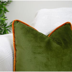 May include: A square throw cushion made of olive green velvet fabric. The cushion features a distinct orange velvet piping trim along its edges, creating a clear contrast against the deep green surface of the soft, plush material.