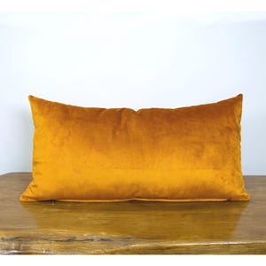 Puede incluir: Un cojín rectangular con una funda de tela de terciopelo de color amarillo mostaza sólido. El cojín tiene una textura suave y lisa con un color consistente en toda la superficie, mostrando un diseño simple con bordes cosidos en todos sus lados.
