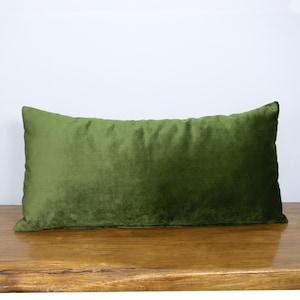 Puede incluir: Un cojín rectangular cubierto con una tela de terciopelo de color verde oliva sólido. El cojín presenta una textura suave y afelpada con un brillo sutil en toda su superficie, mostrando un color verde profundo constante en toda su forma.