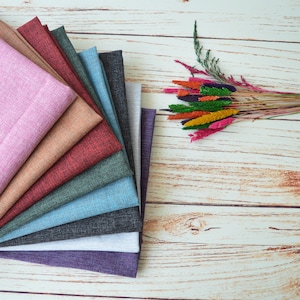 May include: A stack of ten fabric swatches in various colors and textures, including pink, brown, red, green, blue, gray, and purple. The swatches are arranged in a fan shape on a light wood surface. A bouquet of dried flowers with colorful plumes is in the upper right corner of the image.