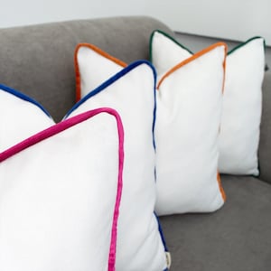 May include: A set of four square white throw pillows, each featuring a distinct colored fabric piping edge. The pillows are finished with magenta, royal blue, orange, and dark green trim, showcasing a textured fabric surface on each cushion.