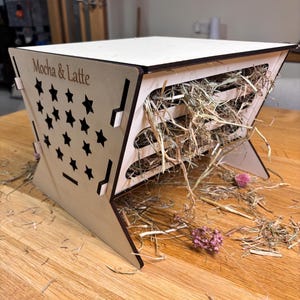 May include: A wooden pet feeder with a star cut-out design and the text "Mocha & Latte". The feeder is filled with hay and dried flowers. The feeder is made of light-colored wood and has a unique angled shape.