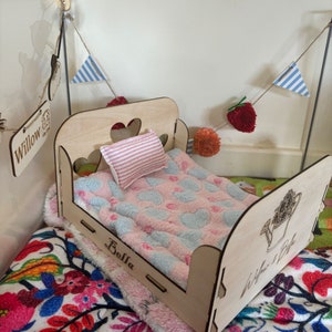 May include: A wooden doll bed with a pink and blue heart patterned blanket and a pink and white striped pillow. The bed is labeled "Bella" and "Willow & Bella".
