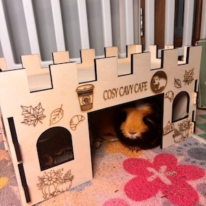 May include: A wooden castle-shaped pet house with laser-cut details, including leaves, a coffee cup, and the text "COSY CAVY CAFE." A guinea pig is inside. The house sits on a floral patterned rug.