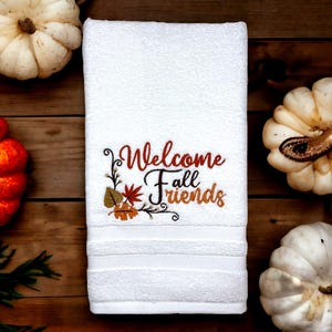May include: White hand towel embroidered with "Welcome Fall Friends" in brown, orange, and black. The design includes fall leaves and decorative flourishes. The towel is surrounded by pumpkins on a wooden surface.