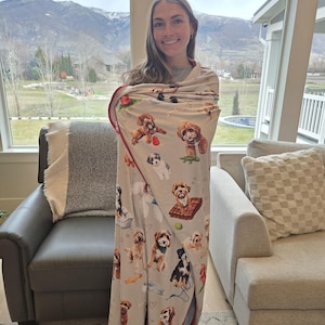 May include: A light gray blanket featuring various dog breeds in playful poses. The blanket has a red border and is wrapped around a person. The background shows a mountain range and a living room setting.