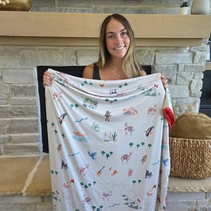 May include: A cream-colored blanket with a red border, featuring a repeating pattern of llamas, cacti, and other Southwestern-themed illustrations. The blanket is held up, showcasing its design and soft texture.