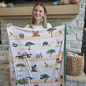 May include: A cream-colored blanket with a green border, featuring a repeating pattern of African wildlife, people, and architecture. The design includes illustrations of birds, trees, and traditional huts. The blanket is held up by a person.