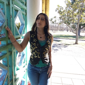 May include: Black vest adorned with gold star-shaped sequins. The vest is sleeveless and has a zipper closure. The person is wearing a teal top and blue jeans. The background includes a turquoise and white structure and a park.