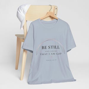 May include: A light blue short-sleeved t-shirt featuring a rose gold arch graphic with the black text BE STILL AND KNOW THAT I AM GOD, a small rose gold heart, and the text PSALM 46:10. A white baseball cap hangs on the wooden hanger behind the shirt.