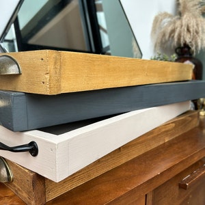May include: Three wooden trays stacked on top of each other. The top tray is natural wood with a gold metal handle. The middle tray is painted dark grey with a gold metal handle. The bottom tray is painted white with a black metal handle. The trays are sitting on a wooden surface.