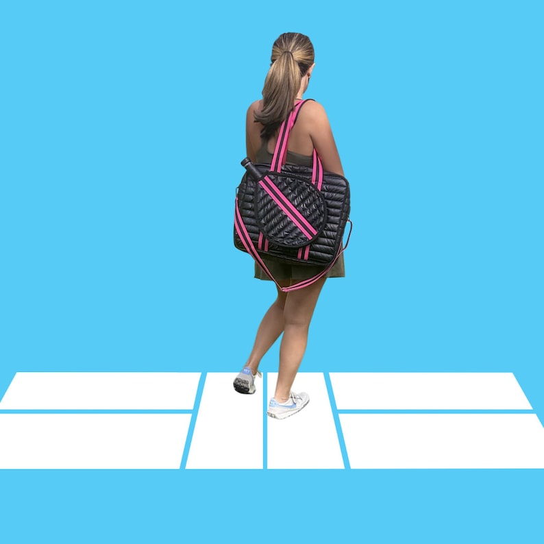 Puede incluir: Una bolsa de tenis acolchada negra con detalles rosas y una raqueta de tenis blanca en su interior. La bolsa la lleva una persona que camina por una pista de tenis blanca.