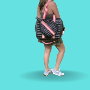 Puede incluir: Una bolsa de tenis acolchada negra con rayas rosas y blancas. La bolsa tiene un soporte para raquetas y un compartimento con cremallera. La bolsa la lleva una persona que lleva un top verde y una camiseta blanca.